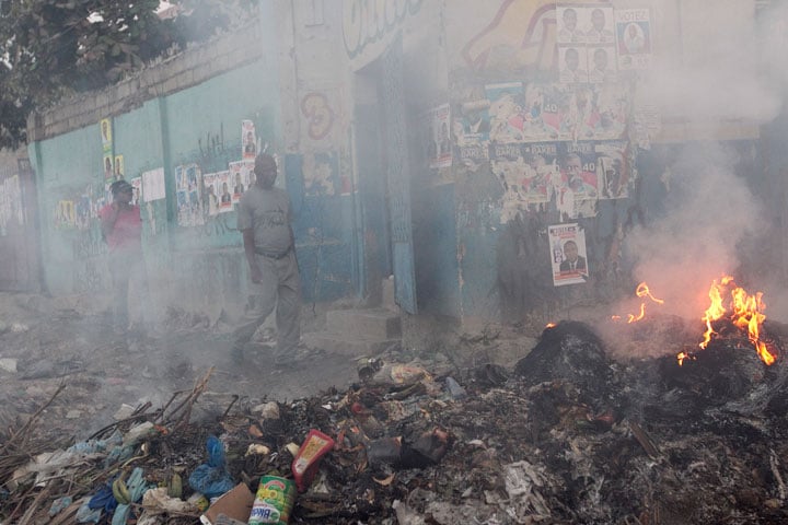 Residents make their way along walls covered with campaign posters and past piles of burning trash in the Carrefour neighborhood of Port-au-Prince.