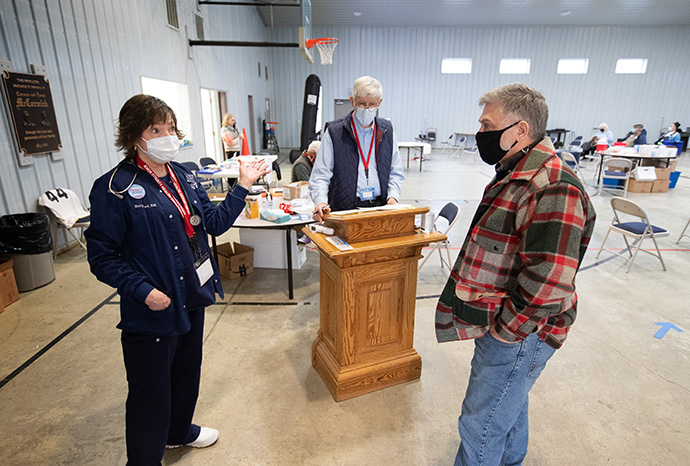 The Rev. Darren Williams (right), pastor of Community United Methodist Church in Vincennes, Ind., visits with Betty Lankford, RN, in the church’s recreational center. The church is hosting a COVID-19 vaccination site, sharing space normally used for basketball games, church meals and a food pantry with local emergency management officials. Lankford is lead nurse for the Knox County (Ind.) Health Department’s COVID-19 response. At the check-in station (center) is volunteer Larry Marchino. Photo by Mike DuBose, UM News.